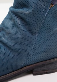 Botte en cuir perforé bleu avec coutures apparentes et semelle en bois sombre et usée.