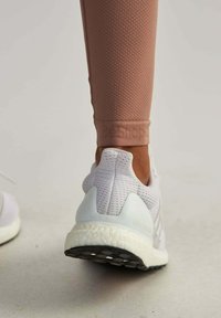 Białe sportowe buty z siateczkowej cholewki, teksturowane beżowe legginsy i czarna gumowa podeszwa, z logo "ultraboost" na pięcie.