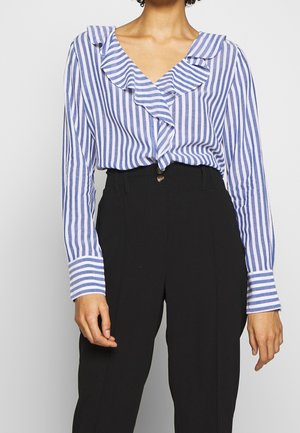 Donna che indossa una camicetta a balze con righe verticali blu e bianche infilata in pantaloni neri a vita alta con bottoni visibili.