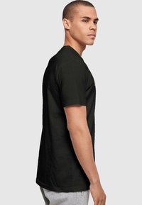 Black cotton t-shirt with short sleeves, crew neck, and a graphic detail on the front. Model wearing grey sweatpants, standing sideways.