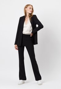 Schwarzer maßgeschneiderter Blazer über einer cremefarbenen V-Ausschnitt-Bluse, kombiniert mit schwarzen Schlaghosen und weißen Sneakers. Glatter Stoff, elegantes Design.