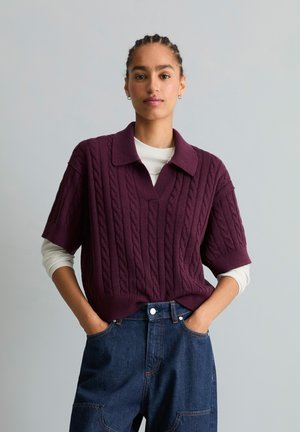 Femme aux cheveux tressés portant un pull violet à manches courtes en maille torsadée par-dessus une chemise blanche à manches longues et un jean bleu, les mains dans les poches.