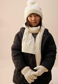 Balts adīts beanie ar pom-pomu, adīta kaklasaite un cimdi, viss apvienots ar melnu pūkainu jaku virs svītraina krekla.