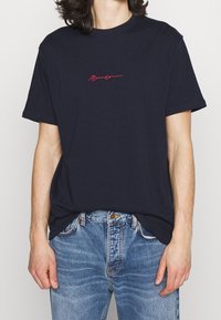 Mörkblå bomullströja med korta ärmar, med en liten röd kursiv logotyp på bröstet. Bärs med ljusblå denimjeans.