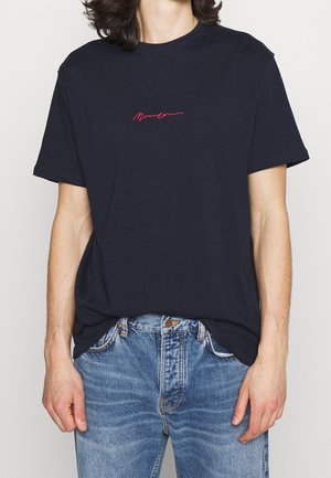Navyblaue Baumwoll-T-Shirt mit kurzen Ärmeln, das ein kleines rotes, kursives Logo auf der Brust trägt. Getragen mit hellblauen Jeans.