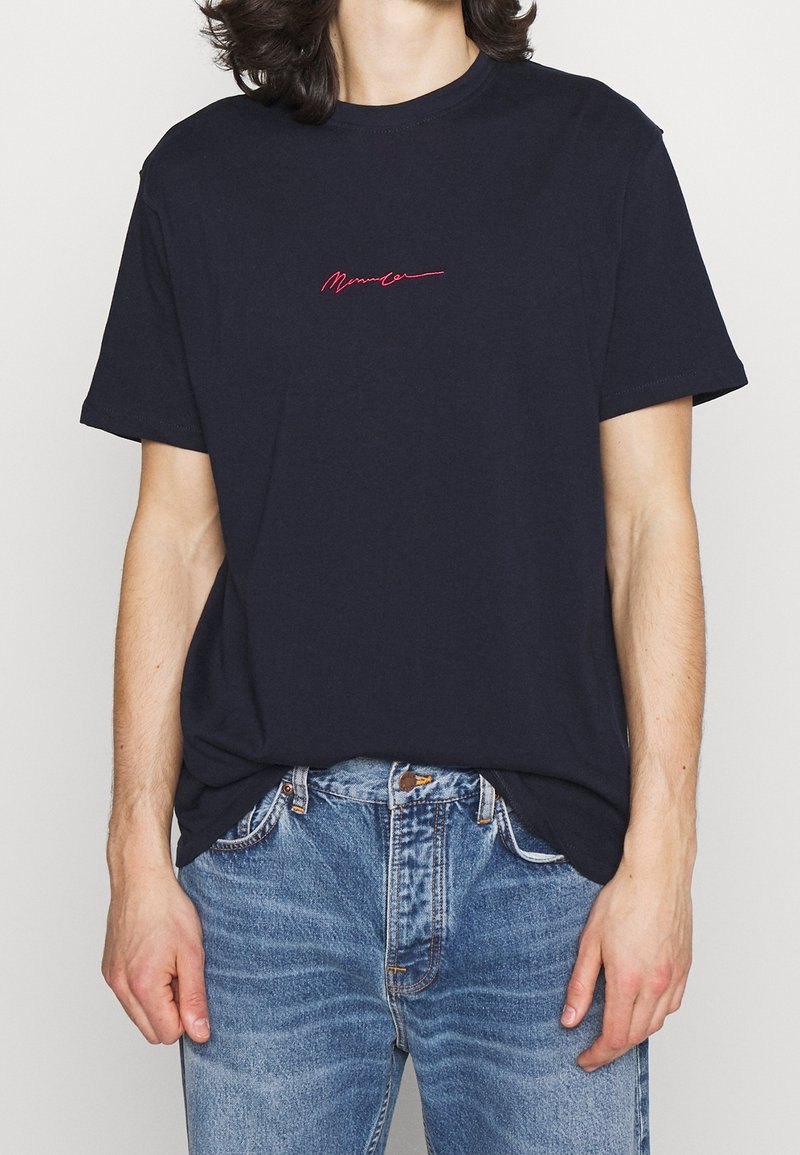 Mörkblå bomullströja med korta ärmar, med en liten röd kursiv logotyp på bröstet. Bärs med ljusblå denimjeans.