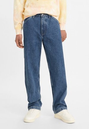 Personne portant un jean en denim bleu ample, des baskets blanches et un sweat-shirt tie-dye jaune clair, debout devant un fond uni.