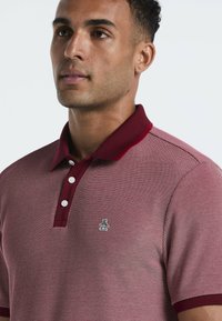 Polo shirt rayé rouge et blanc avec un col bordeaux, comportant trois boutons blancs et un petit logo de pingouin sur la partie gauche de la poitrine.