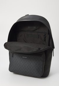 Zaino nero con un esterno in pelle liscia e una tasca frontale a motivo geometrico. Presenta un vano principale con zip e tasche interne.