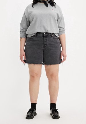 Persona que lleva una camisa gris de manga larga, pantalones cortos vaqueros negros con bordes deshilachados, calcetines negros y zapatos negros brillantes con cordones.