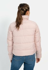 Lys pink pufferjakke med høj krave, horisontal syning og en cropped design. Bæres over blå jeans, glat tekstur.