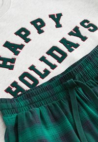 Huppari harmaa, jossa on punaisella ja vihreällä brodeerattu "HAPPY HOLIDAYS", yhdistettynä vihreisiin ja laivastonsinisiin ruudullisiin flanellihousuihin, joissa on joustava vyötärö.