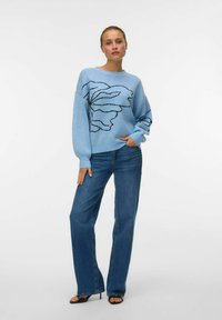 Svijetloplavi pleteni pulover s crnim cvjetnim uzorkom, uparen s tamnoplavim trapericama visokog struka. Ima duge rukave i opušteni kroj.