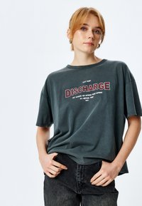 Czarny t-shirt z bawełny w ciemnoszarym kolorze, o luźnym kroju, z krótkim rękawem i okrągłym dekoltem. Na przodzie znajduje się grafika "DISCHARGE" w czerwonym i białym kolorze.