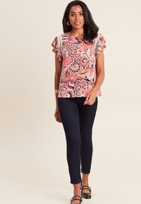 Kortebloezige, roze paisley blouse met ruches mouwen, gecombineerd met navy slim-fit jeans en zwarte sandalen met banden. Gladde textuur en levendig patroon.