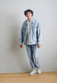 Denim jacket in light blue with front pockets, layered over a white shirt, paired with light blue jeans and white sneakers. Minimalist design.