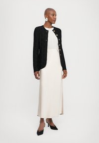 Black textured cardigan with gold buttons, layered over a light grey top and beige skirt. Paired with black pointed-toe heels.