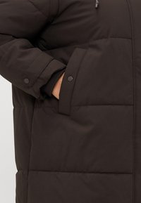Brown insulated jacket featuring a quilted design, a side pocket with a flap, and a soft texture. The fabric is smooth with a matte finish.