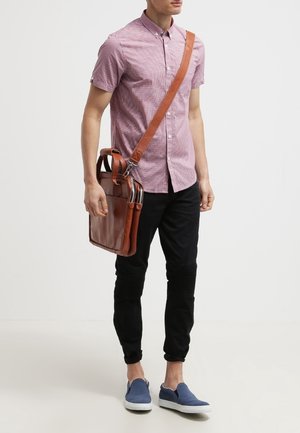 Hombre lleva una camisa de cuadros roja de manga corta, pantalones negros, zapatos azules sin cordones, y lleva un bolso de cuero marrón al hombro con bolsillos delanteros y cremalleras.