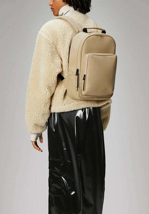 Zaino beige con una texture liscia, forma rettangolare, tasca frontale con zip e spallacci regolabili, indossato su una giacca di pile crema e pantaloni neri lucidi.