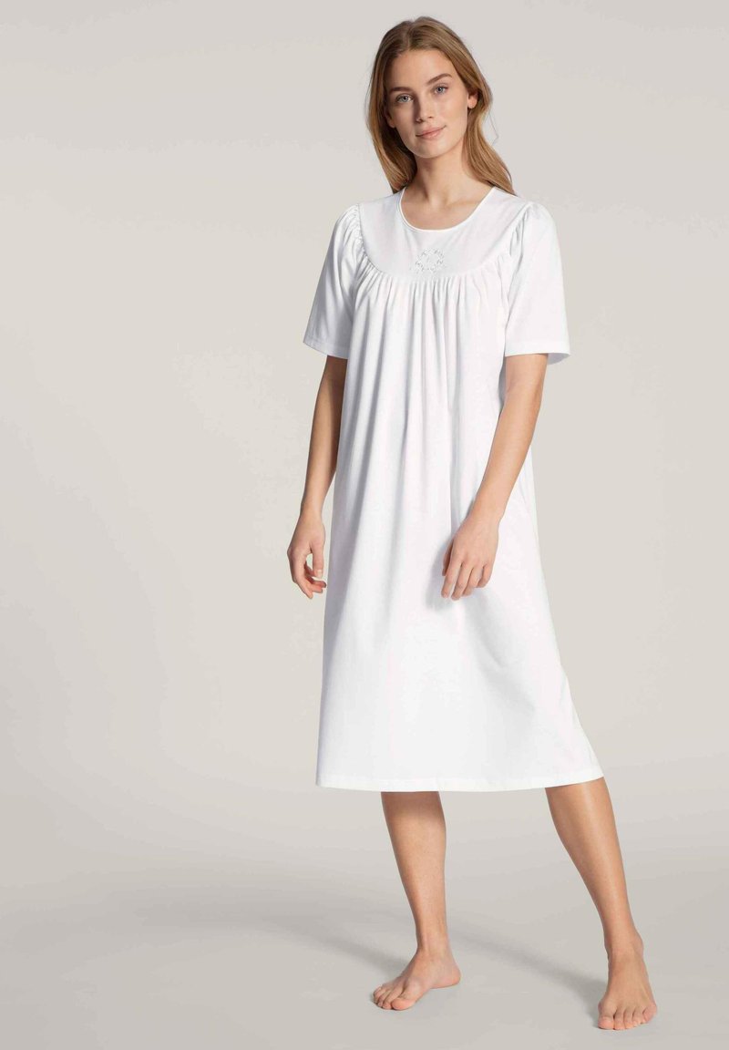 Camicia da notte bianca in cotone con maniche corte, scollatura rotonda e dettagli arricciati sotto il seno, che scende sotto il ginocchio. Tessuto a trama semplice.