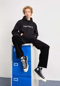 Svart hettegenser med hvit logo, svarte kordfløyelsbukser og svarte høy-top sneakers. Modellen sitter på en blå arkivskap.