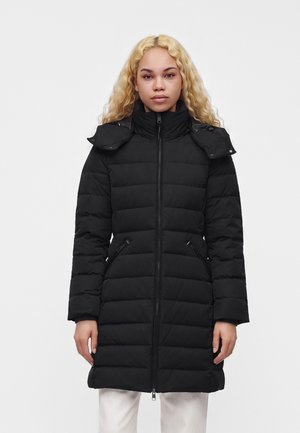 Femme aux longs cheveux blonds bouclés portant un manteau noir matelassé zippé avec un col montant et des poches latérales zippées.