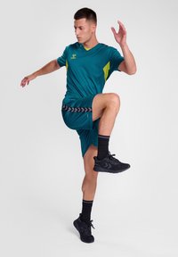 Teal idrottskläder bestående av en kortärmad topp med gula detaljer och matchande shorts. Svarta sneakers med texturerade sulor.