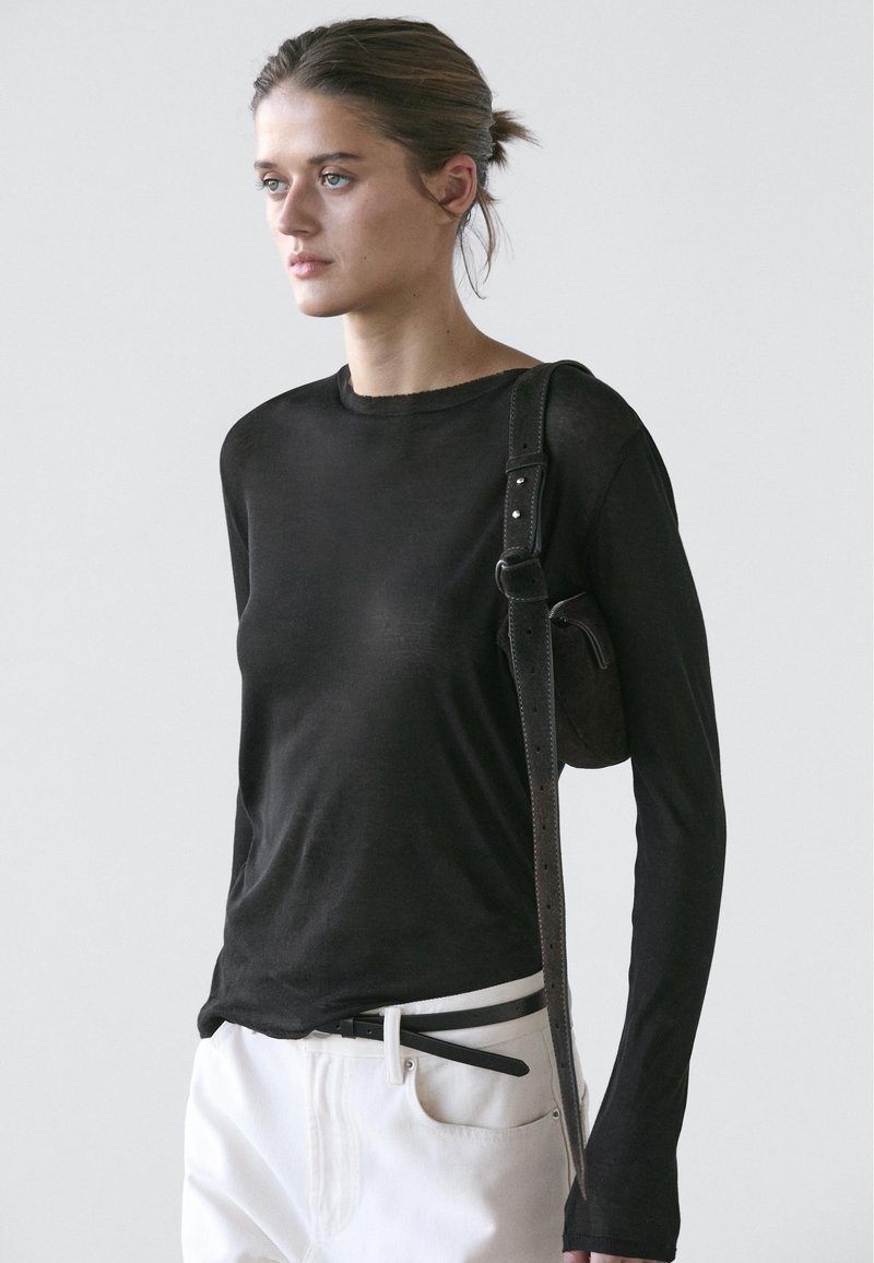 Young woman with pulled-back hair wearing a fitted black long-sleeve top, white pants with a black belt, and a black shoulder bag.