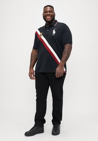 Črna polo majica z rdečo in belo diagonalno črto, logotip na prsih, v kombinaciji s črnimi hlačami in črnimi supergami. Model stoji in se smeška.