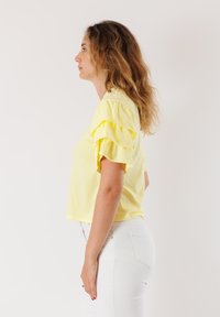 T-shirt giallo corto con maniche arricciate, realizzato in tessuto morbido. Indossato con jeans bianchi a vita alta, che mostrano il profilo laterale dell'outfit.