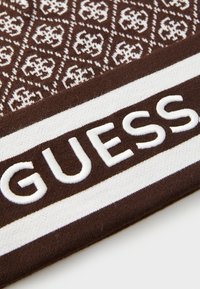 Brunt tekstil med et hvitt geometrisk mønster og en fremtredende brodert hvit "GUESS"-etikett på en kontrasterende stripet bakgrunn.
