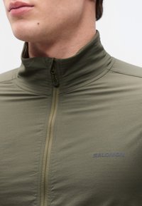 Ανδρας που φοράει ελιάς νερουλί ζακέτα με φερμουάρ και ψηλό γιακά, με το λογότυπο Salomon στο στήθος, εμφανιζόμενος από τον λαιμό μέχρι τον άνω κορμό.