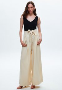 Lichtbeige wijde pantalon met een stoffen riem, hoge taille, plooien aan de voorkant en zijzakken, gecombineerd met een zwarte aansluitende tanktop.