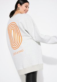 Felpa grigia chiara con grafica ovale concentriche arancioni e testo "COOL & KIND" sul retro. Caratterizzata da una vestibilità comoda e orlo a coste.