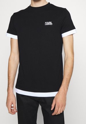Una persona que lleva una camiseta negra de Karl Lagerfeld con ribete blanco en las mangas y el dobladillo, de pie frente a un fondo liso.