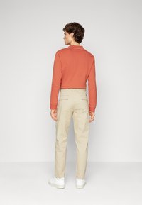 Maglia a maniche lunghe color ruggine con scollo rotondo, abbinata a pantaloni beige con tasche posteriori, mostrata da dietro. Sneakers bianche visibili.