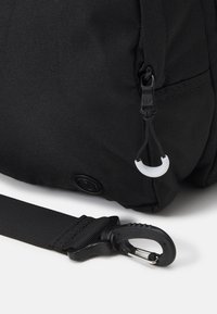 Czarny plecak z tkaniny o teksturowanej powierzchni, z suwakiem i klipsowym zapięciem na pasku. Udoskonalony okrągłą metką.