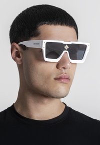 Hvide oversized solbriller med et geometrisk design, mørke linser og guldaccentuer på rammen. En simpel sort t-shirt bæres.