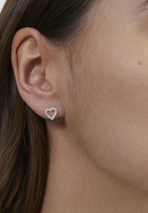 Close-up of a woman's ear wearing a small, silver heart-shaped stud earring with embedded clear stones.