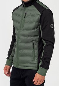 Zielona pikowana kurtka z czarnymi rękawami, pełnym zamkiem, kieszeniami bocznymi, ściągaczami na mankietach oraz naszywką z logo na lewym ramieniu. Różnorodna faktura materiału podkreśla poszczególne części.