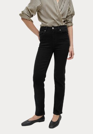 Persoon die zwarte jeans met hoge taille draagt, een beige en zwarte gestreepte blouse met lange mouwen waarvan de mouwen zijn opgerold, en zwarte platte schoenen, staand met ontspannen handen.