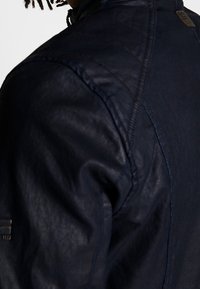 Námořnická modrá kožená bunda s prošívaným designem, texturovaným povrchem a jemným brandingem na rameni. Detail rukávu v detailu.