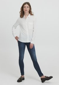 Hvid skjorte med lange ærmer og en brystlomme, parret med mørkeblå skinny jeans og sorte loafers med en guldkæde-detalje.