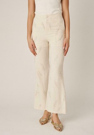 Mujer con pantalones de tiro alto color crema con patrones de trigo bordados y tacones dorados abiertos en los dedos, posando frente a un fondo liso.