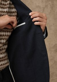 Blazer in lana blu con foderatura interna texturizzata. Presenta una tasca nascosta con un bordo bianco a contrasto, evidenziando dettagli su misura.