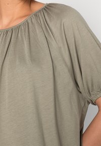 Top grigio leggero con scollatura arricciata e maniche corte a sbuffo. Il tessuto è morbido con una sottile texture.