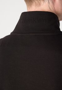 Crna polo majica s visokim ovratnikom, s diskretnim utisnutim logom "CalvinKlein" na dekolteu. Mekan, glatki osjećaj tkanine.