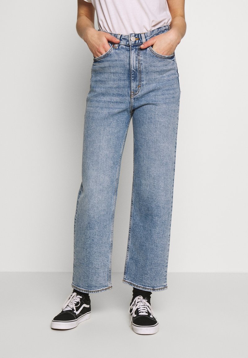 Personne portant un jean large bleu clair, des baskets noires et blanches, un t-shirt blanc, debout avec les mains dans les poches avant.