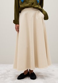 Jupe en coton beige, longue et fluide avec un ourlet à franges. Associée à une chemise vert foncé avec des accents brodés en bleu. Chaussures noires visibles.